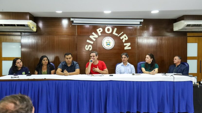 Candidato sentado à mesa com sindicalistas durante reunião, em sala com bandeiras ao fundo