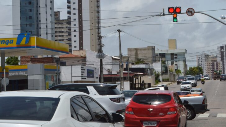 Semáforo em avenida movimentada de Natal, com carros parados na faixa e prédios ao fundo