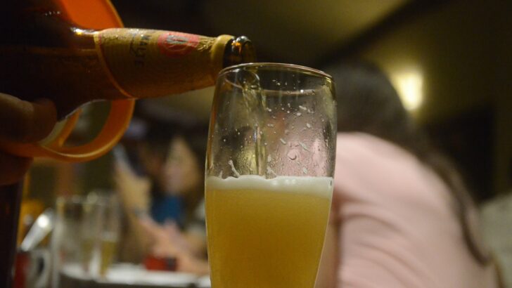 Garçom serve cerveja em taça sobre mesa de bar, com clientes ao fundo