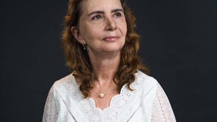 Mulher de blusa branca rendada sentada, com as mãos apoiadas no colo, em retrato sobre fundo escuro
