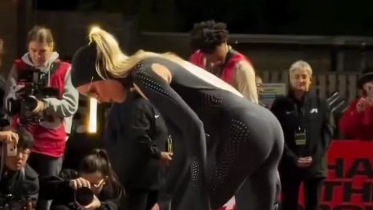 Atleta de macacão preto de corpo inteiro, com detalhes perfurados, inclinada na pista logo após a prova, cercada de fotógrafos