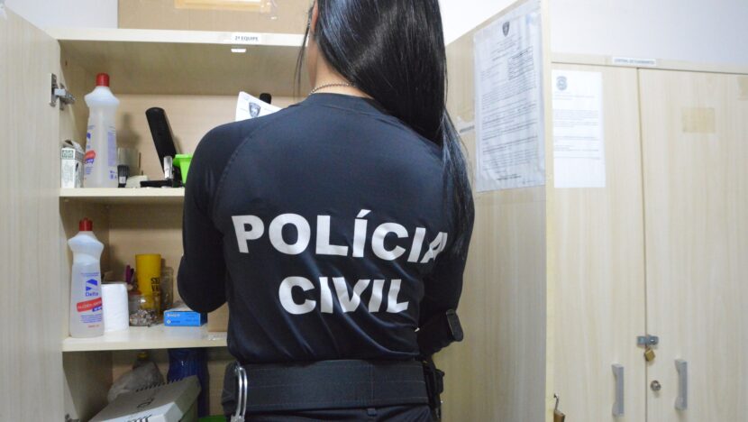 Agente de colete com a inscrição Polícia Civil, de costas, revistando um armário durante diligência