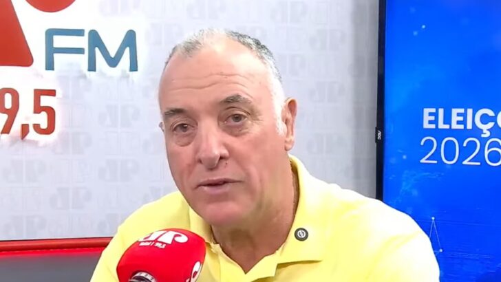 Homem de camisa amarela fala ao microfone em estúdio de rádio, com painel de eleições ao fundo