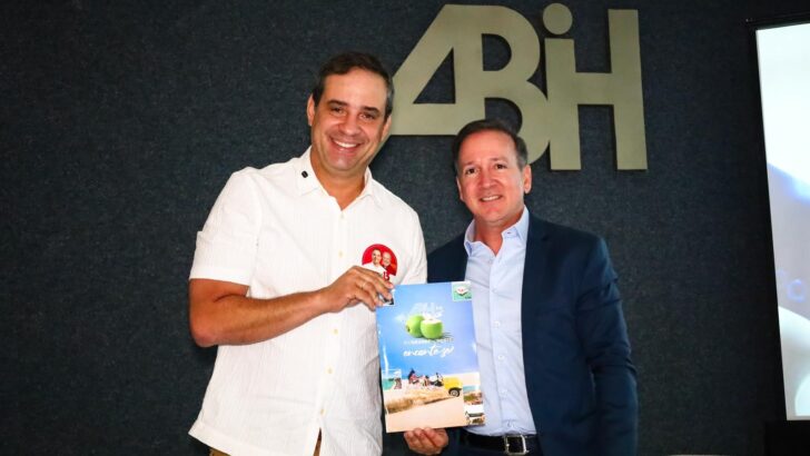 Dois homens em pé diante do letreiro da ABIH; o da esquerda, de camisa branca, segura um exemplar impresso da Agenda da Hotelaria Potiguar