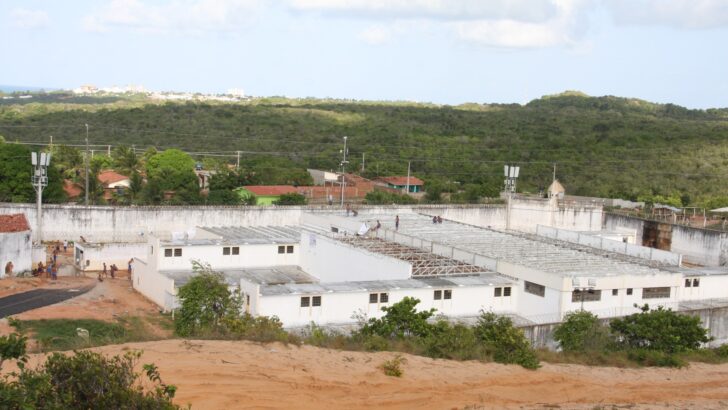 Vista elevada de um complexo penitenciário de pavilhões brancos cercado por muros, com vegetação e colinas ao fundo