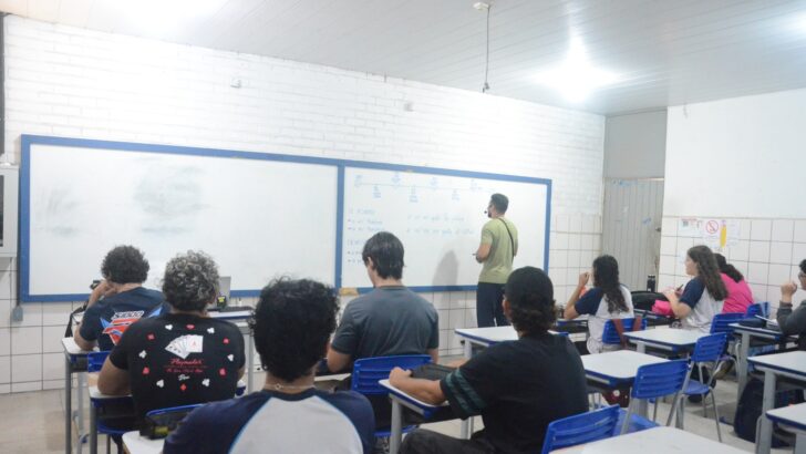 Alunos sentados em carteiras azuis, vistos de costas, durante aula em sala de escola da rede pública, com o professor junto ao quadro branco