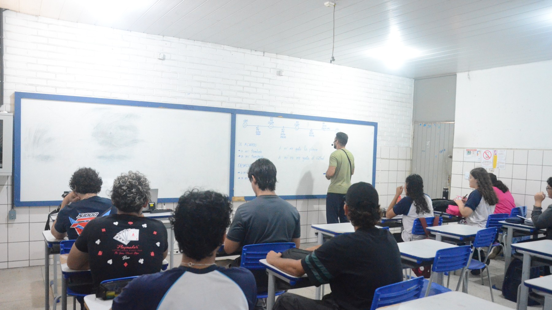 Estudantes de costas em carteiras azuis e professor diante do quadro em sala de aula da rede estadual