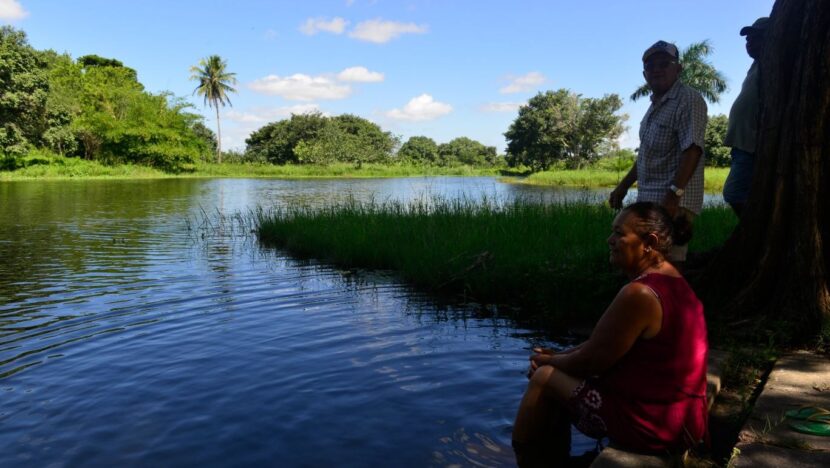 Lagoa de água escura cercada por vegetação e coqueiros, com pessoas na margem, uma delas sentada em primeiro plano