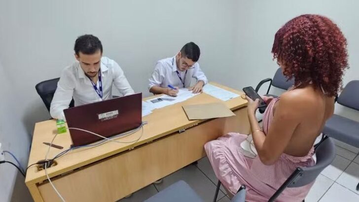 Dois atendentes de camisa branca sentados atrás de uma mesa com notebook e documentos, diante de uma pessoa atendida, vista de costas