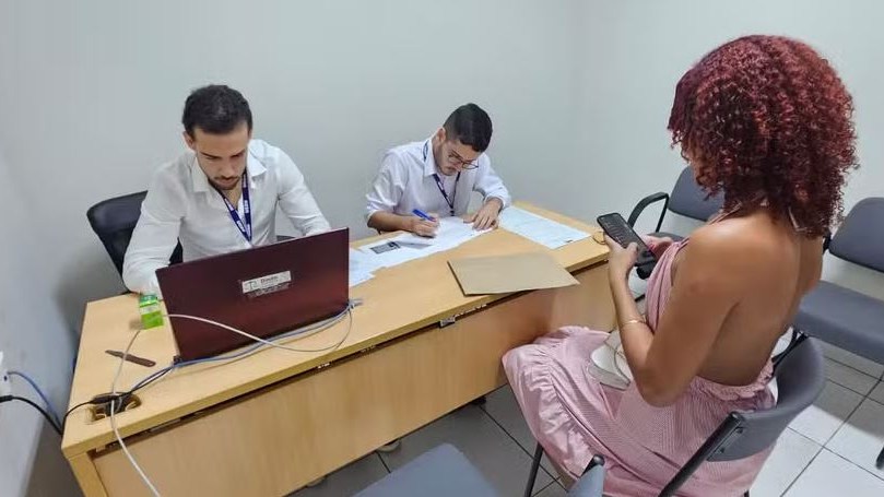 Dois atendentes de camisa branca sentados atrás de uma mesa com notebook e documentos, diante de uma pessoa atendida, vista de costas
