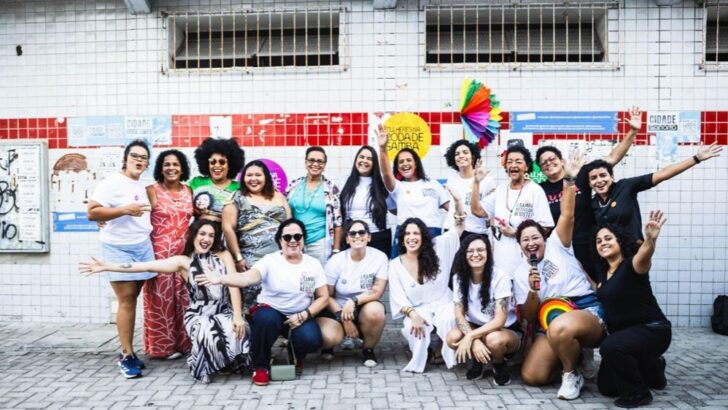 Cerca de vinte mulheres posam juntas diante de um muro de azulejos brancos e vermelhos, muitas com camiseta branca do encontro, uma segurando uma placa e outra um leque colorido
