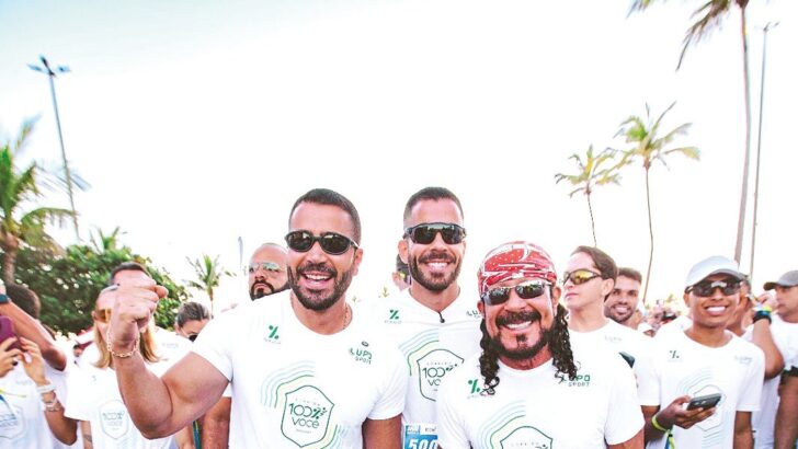 Bell Marques, de bandana vermelha, caminha entre corredores de camiseta branca da Corrida 100% Você, com coqueiros e céu claro ao fundo