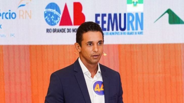 Homem de terno azul e camisa branca fala ao microfone diante de painel com logomarcas de OAB-RN e Femurn