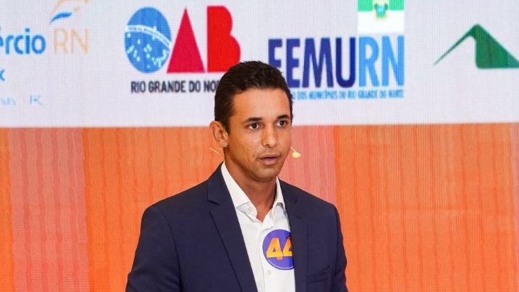 Homem de terno azul e camisa branca fala ao microfone diante de painel com logomarcas de OAB-RN e Femurn