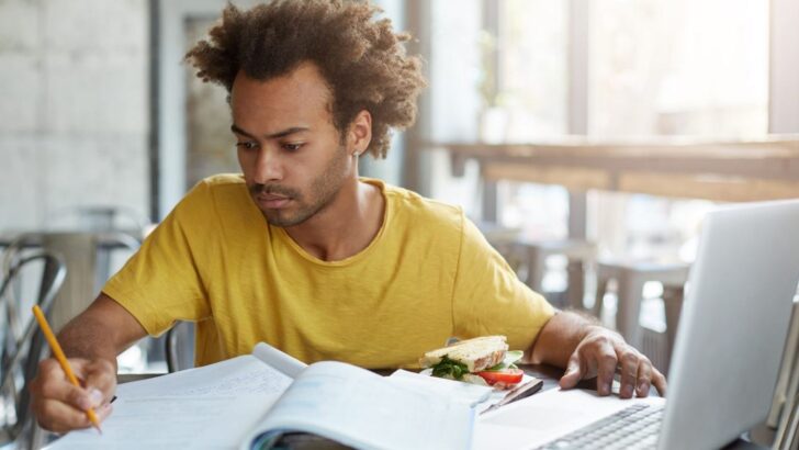 Pessoa estuda em frente ao computador, com caderno e livro abertos sobre a mesa