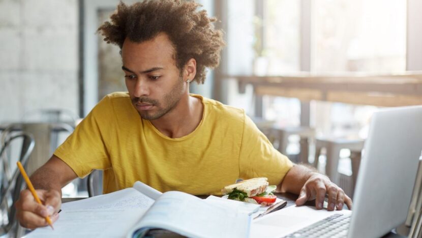 Pessoa estuda em frente ao computador, com caderno e livro abertos sobre a mesa