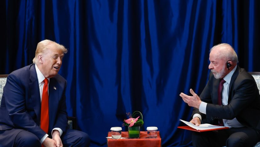 Dois homens de terno sentados em poltronas conversam diante de cortina azul