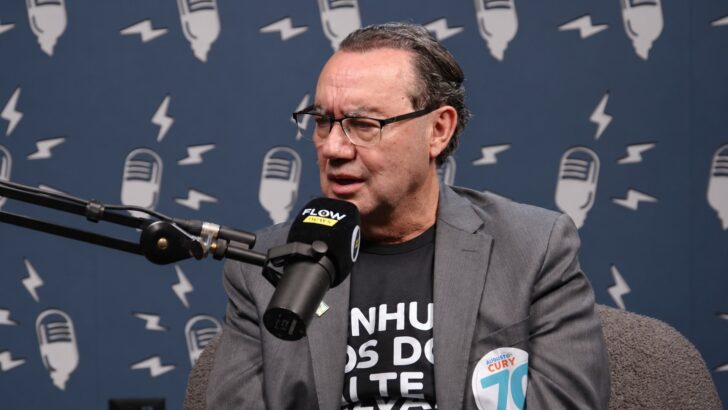 Homem de óculos, paletó cinza e camiseta preta fala ao microfone em estúdio de podcast