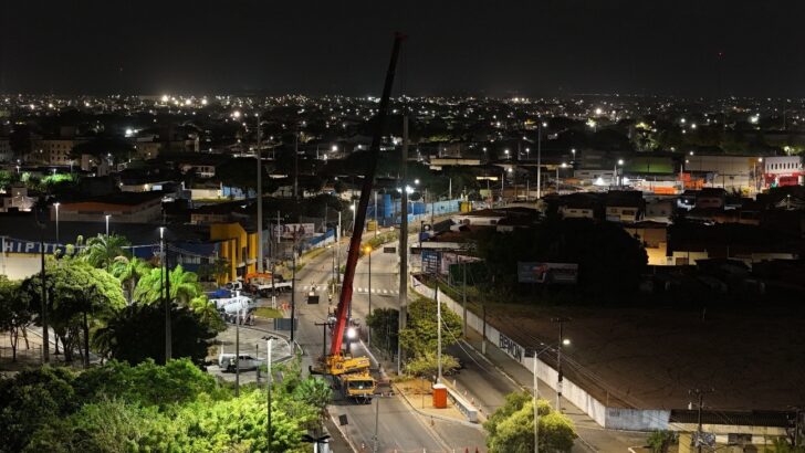 Vista noturna de avenida com guindaste e postes em obra, com a cidade iluminada ao fundo