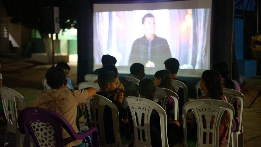 Público sentado em cadeiras plásticas assiste a filme projetado em telão à noite