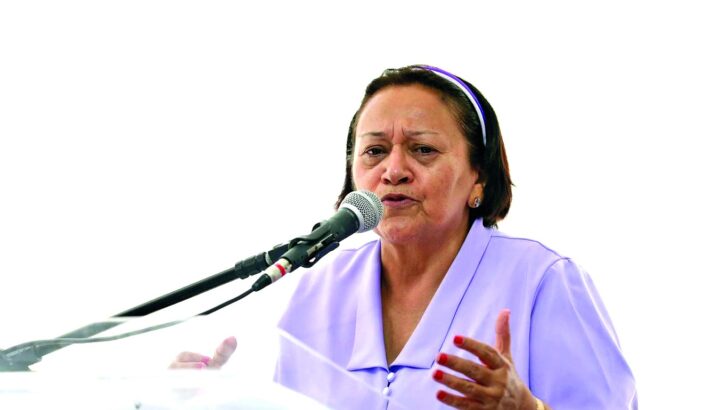 Mulher de blusa lilás fala ao microfone atrás de púlpito