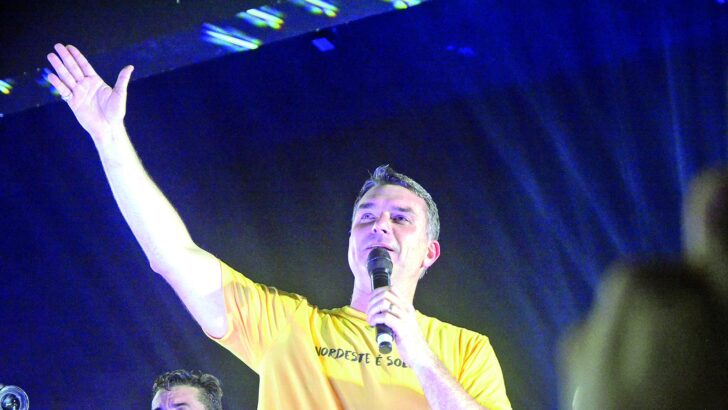 Homem de camiseta amarela ergue o braço e fala ao microfone em palco