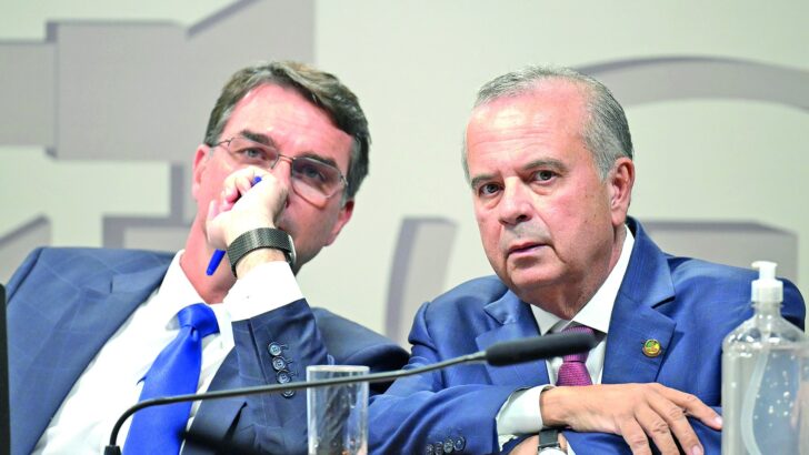 Dois homens de terno sentados à mesa diante de microfones