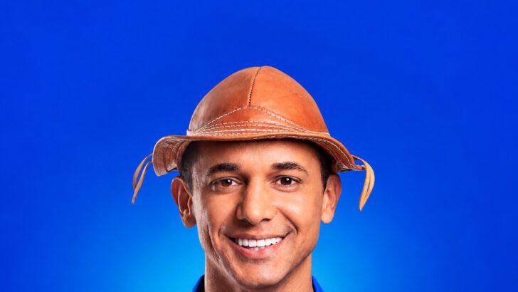 Homem sorridente de chapéu de couro e camisa azul diante de fundo azul