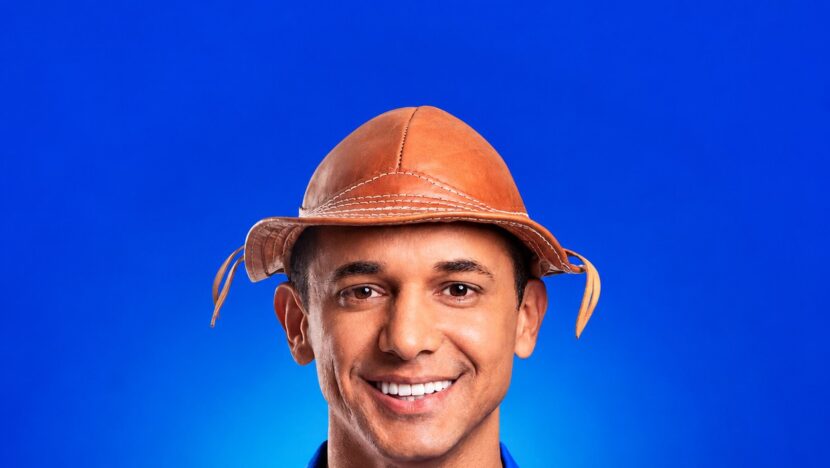 Homem sorridente de chapéu de couro e camisa azul diante de fundo azul