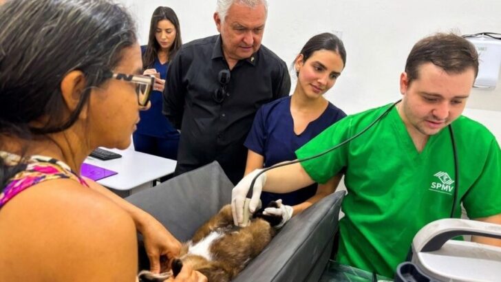 Veterinário de jaleco verde examina um cão em mesa de atendimento, observado por General Girão e outras pessoas