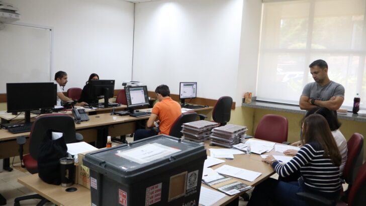 Urna eletrônica em caixa sobre mesa de escritório, com servidores trabalhando ao fundo
