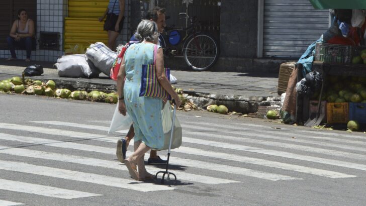 Mulher idosa com bengala atravessa faixa de pedestres em rua de comércio