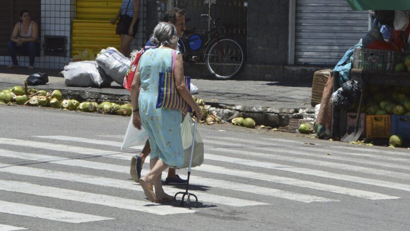 Mulher idosa com bengala atravessa faixa de pedestres em rua de comércio