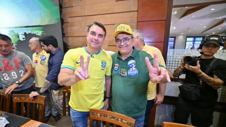 Dois homens, um de camisa amarela da seleção e outro de camisa verde e boné amarelo, fazem sinal de vitória com as mãos em restaurante