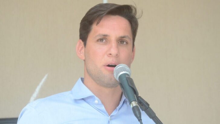 Homem de cabelo escuro e camisa azul-clara fala ao microfone