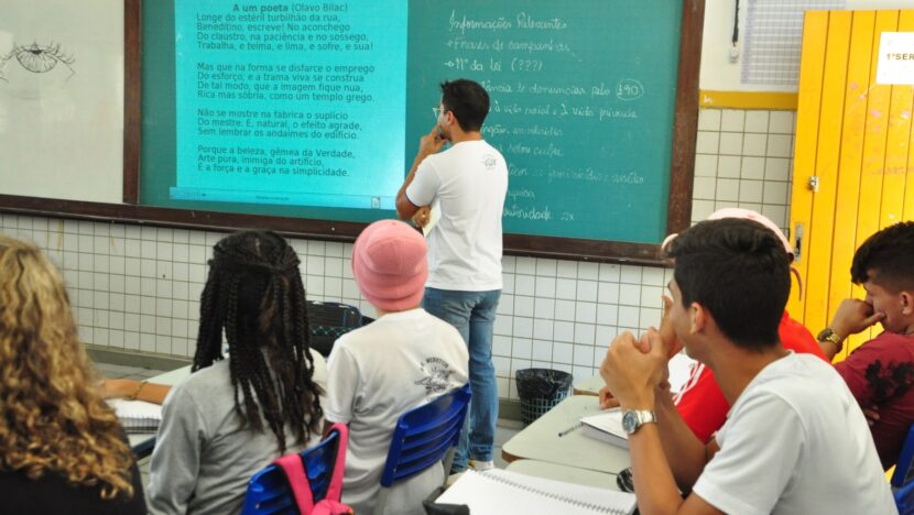 Estudantes sentados em sala de aula observam professor diante de quadro verde e projeção com um poema