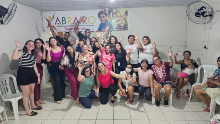 Grupo de mulheres sorridentes acena para a câmera em sala com cadeiras brancas e banner ao fundo