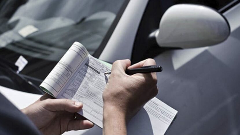 Mão preenche talão de multa de trânsito com caneta ao lado do retrovisor de um carro