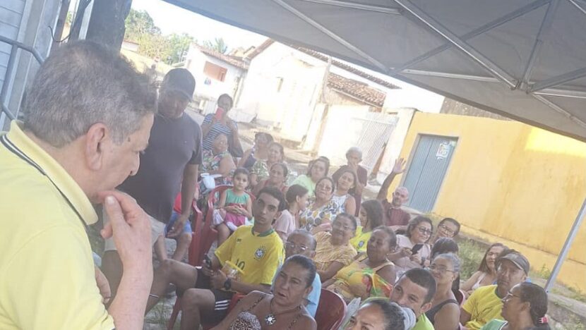 Homem de camisa amarela, de costas, fala a moradores sentados em cadeiras sob uma tenda