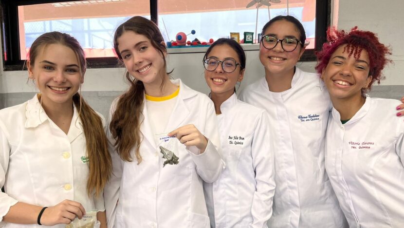 Cinco jovens de jaleco branco sorriem lado a lado em laboratório escolar
