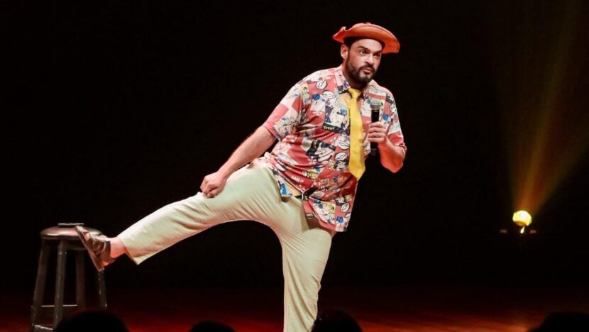 Humorista de chapéu laranja, camisa estampada e gravata amarela ergue a perna no palco com microfone na mão