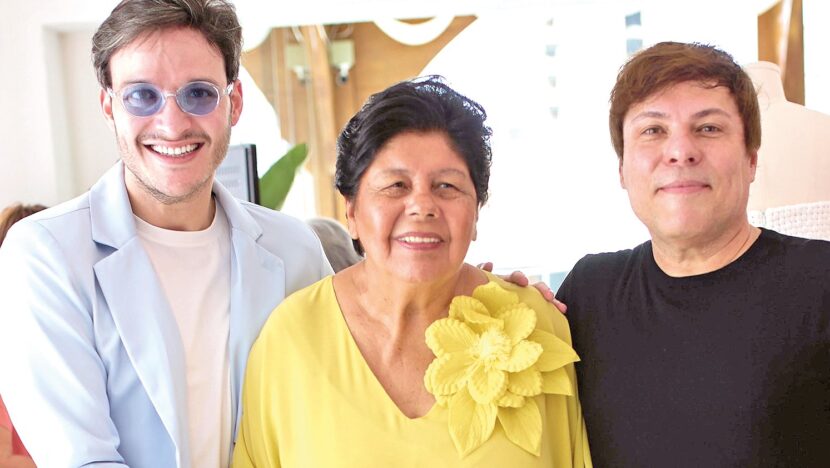 Dois homens e uma mulher de blusa amarela com flor sorriem lado a lado