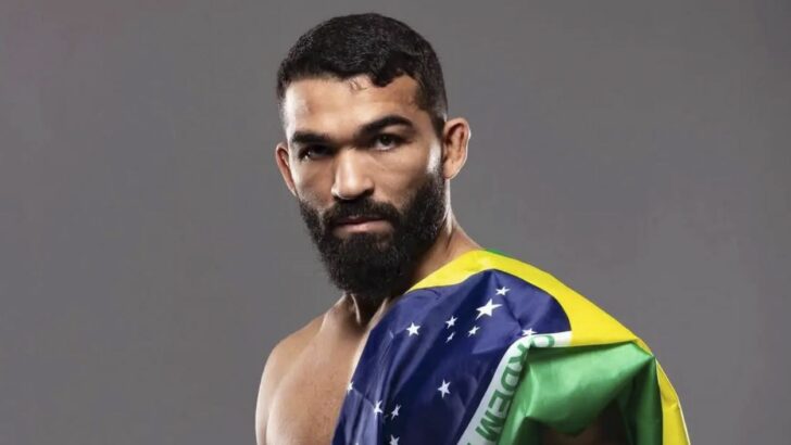 Lutador de barba, sem camisa, com a bandeira do Brasil sobre o ombro, diante de fundo cinza