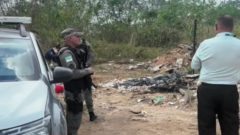 Policial militar ao lado de uma viatura e outras pessoas em uma estrada de terra cercada por vegetação e entulho