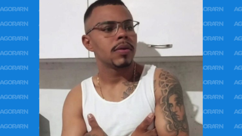 Homem de óculos, camiseta branca e braços cruzados em ambiente interno