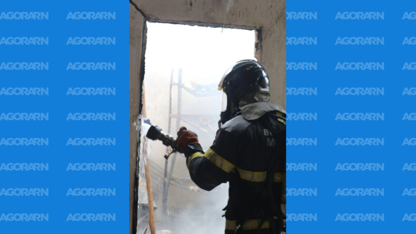 Bombeiro equipado com capacete e roupa de proteção utiliza mangueira durante combate a incêndio em imóvel