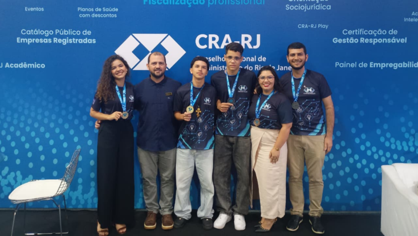 Grupo de estudantes e adultos posa para foto diante de painel do Conselho Regional de Administração do Rio de Janeiro, com participantes exibindo medalhas