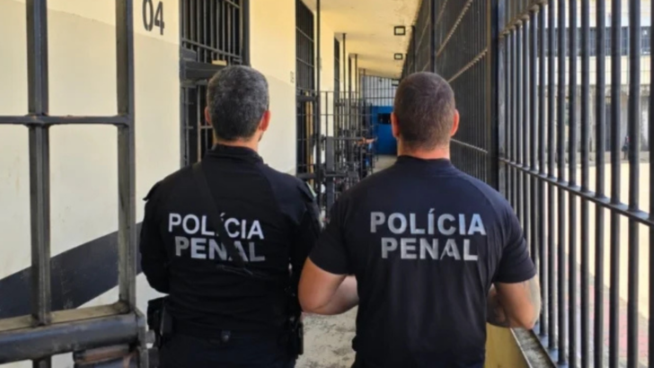 Dois policiais penais vistos de costas em corredor de unidade prisional