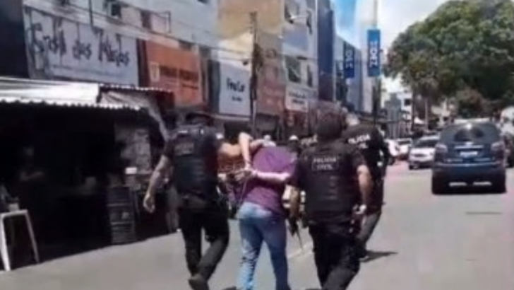 Dois agentes conduzem um homem por uma rua durante operação policial em Natal.