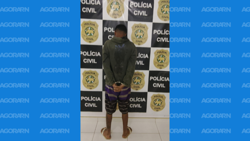 Homem de costas e com as mãos algemadas diante de painel da Polícia Civil do Rio Grande do Norte